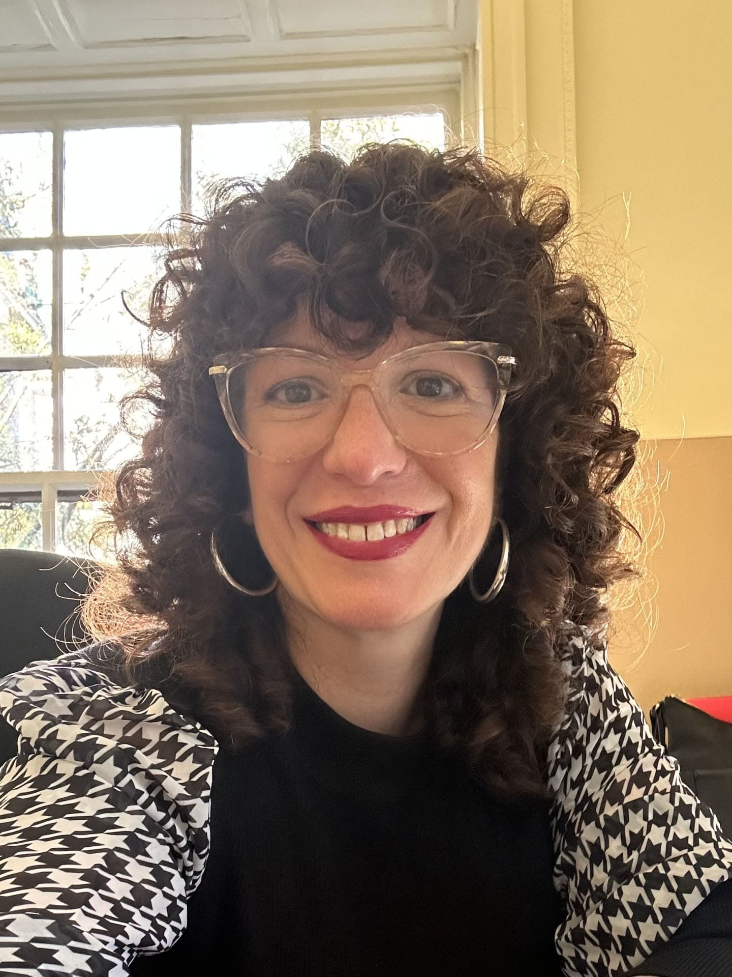 Smiling woman with curly, dark hair, glasses, and hoop earrings, wearing a houndstooth scarf.